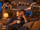 Man relaxing on a sofa beneath a miniature train looping through repeated workplace conversations, illustrating overthinking a conversation.